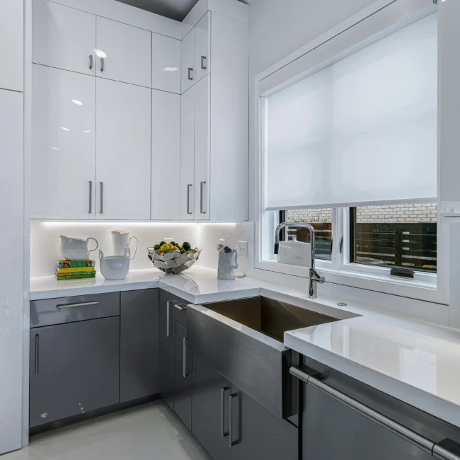 White kitchen with farmhouse sink, gray lower cabinets and roller shade in Urban Oasis by Mike Stevens Homes