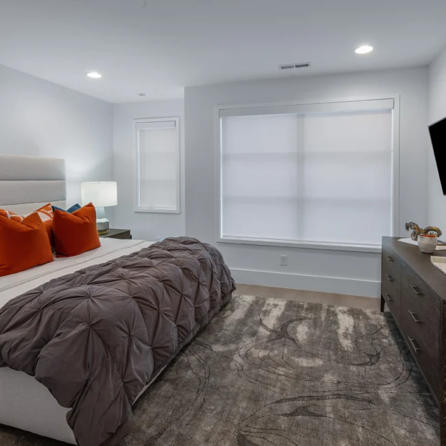 Bedroom with orange throw pillows, gray bedding and roller shade in Urban Oasis by Mike Stevens Homes