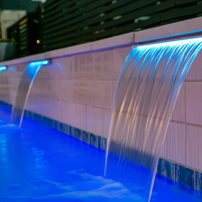 Illuminated blue waterfall spout pouring into pool wall in Urban Oasis by Mike Stevens Homes