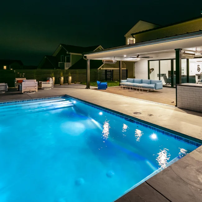 Illuminated pool with stone deck and covered outdoor lounge at night in Urban Oasis by Mike Stevens Homes