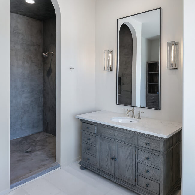 Bath with arched mirror, gray vanity and walk-in shower in Minimalist French Farmhouse, a custom home by Mike Stevens Homes