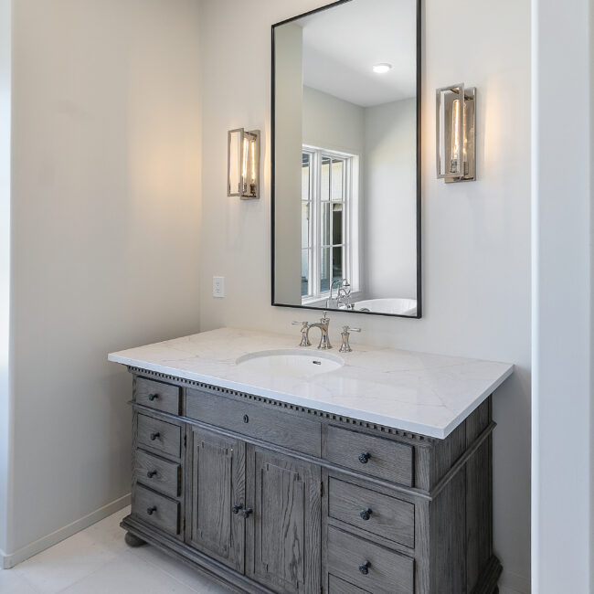 Gray vanity with framed mirror and wall sconces in Minimalist French Farmhouse, a custom home by Mike Stevens Homes