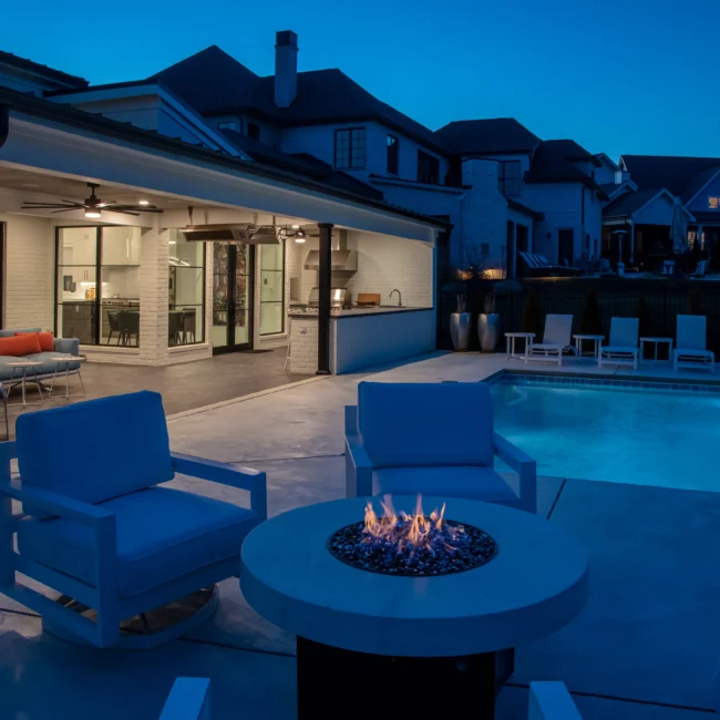 Covered patio at dusk with blue chairs and round fire table in Urban Oasis by Mike Stevens Homes