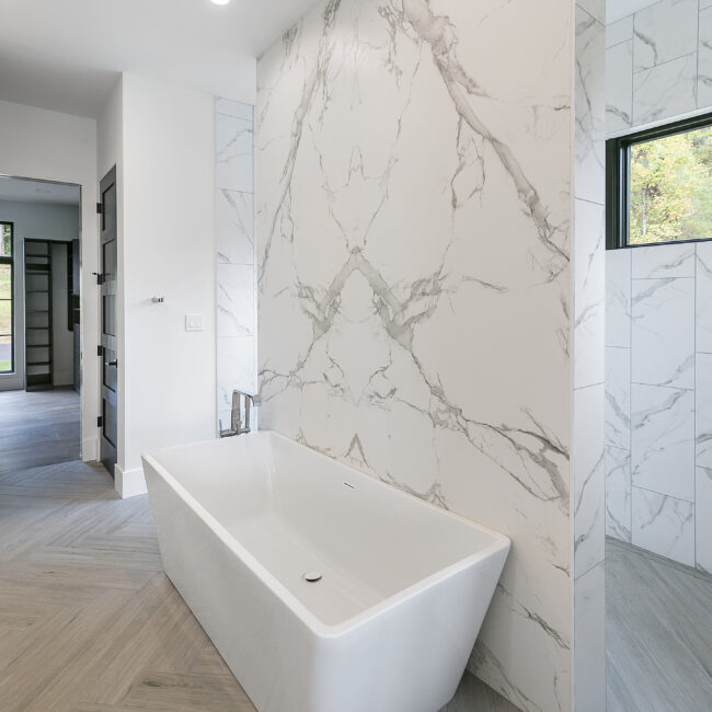Freestanding white tub before marble-look porcelain wall in Rocky Top Hideaway, a custom home by Mike Stevens Homes