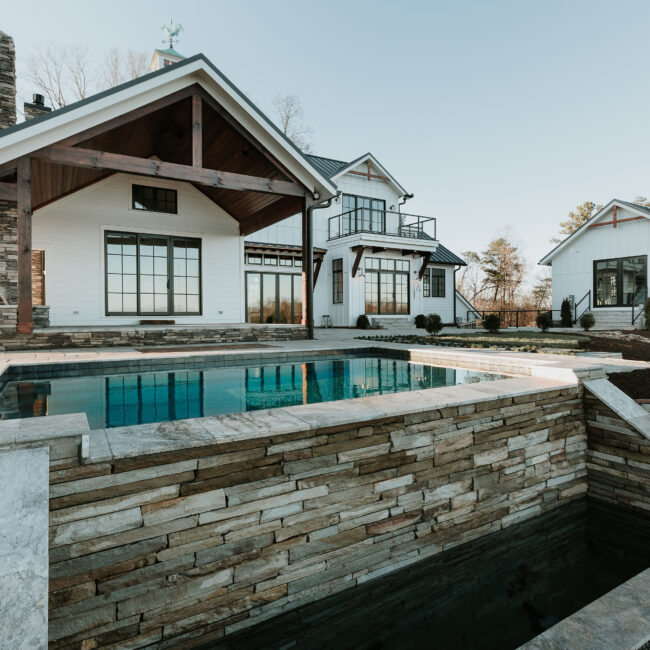 Stone-walled pool beside covered porch of white farmhouse-style home in Mountain Vista by Mike Stevens Homes
