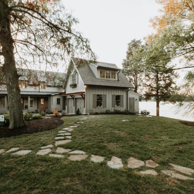 Dormered home with stone pathway and mature shade trees in Island Farmhouse, a custom home by Mike Stevens Homes
