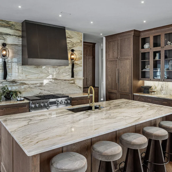 Marble island with gold faucet and bar stools beside walnut glass-front cabinets in The Sanctuary by Mike Stevens Homes