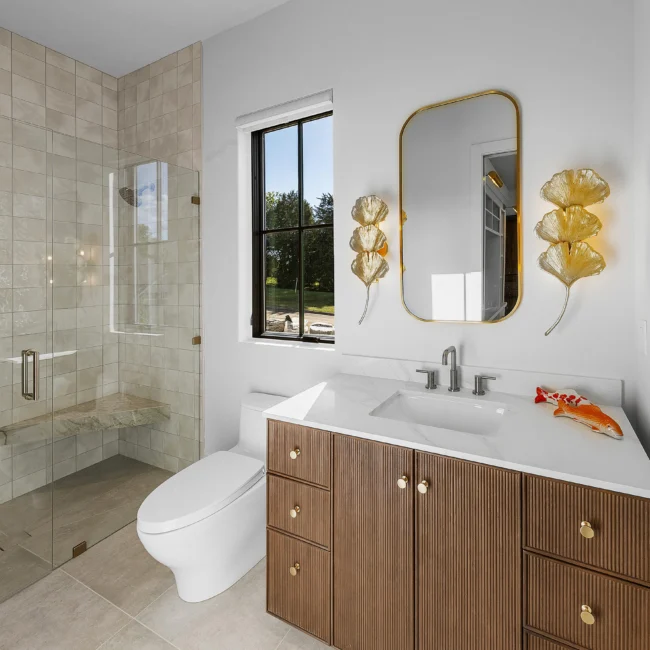 Bathroom with walk-in glass shower, double-sink vanity and round gold mirror in The Sanctuary by Mike Stevens Homes