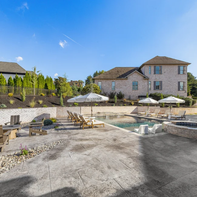 Backyard pool and stone patio with umbrellas and pergola beside brick home in The Sanctuary by Mike Stevens Homes