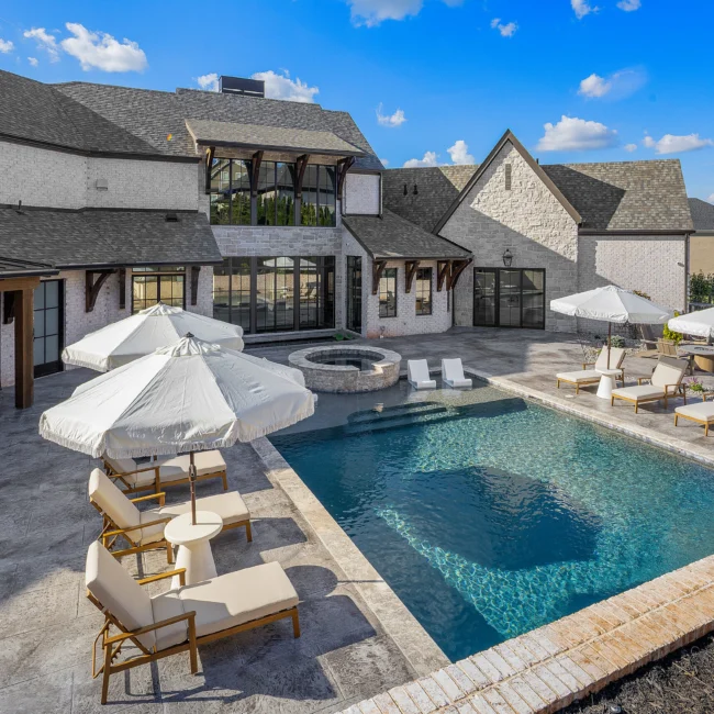 Pool with cream umbrellas, lounge chairs and fire bowl beside rear of stone home in The Sanctuary by Mike Stevens Homes