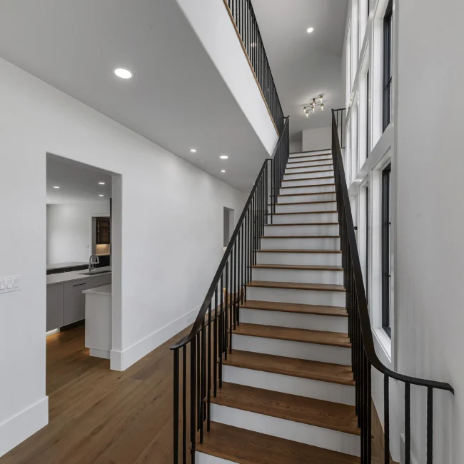 Wood staircase with black rails rising beside a glass-walled hall in Edgewater Estate, a custom home by Mike Stevens Homes