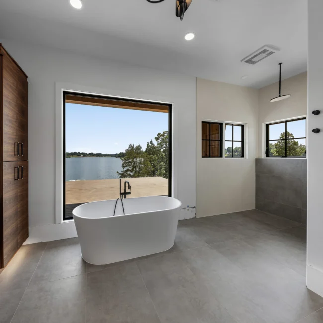 Freestanding tub by a lake-view window beside a walnut linen tower in Edgewater Estate, a custom home by Mike Stevens Homes