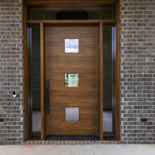 Walnut front door with transom and sidelights between dark brick walls at Modern Prairie by Mike Stevens Homes