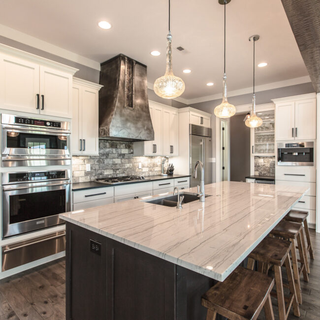 Kitchen with hammered-metal range hood and double wall ovens in Rockport Gray Farmhouse by Mike Stevens Homes