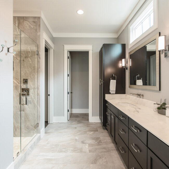 Bath with dark double vanity and glass tile shower in Rockport Gray Farmhouse by Mike Stevens Homes