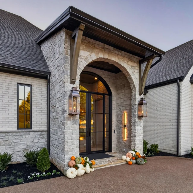 Sunset Heights arched stone entry with black steel-framed double doors, brass lanterns and pumpkins on the steps, flanked by white brick walls