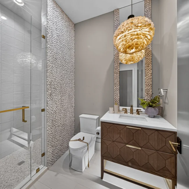 Bathroom with a pebble-tile glass shower, brass-trimmed dark vanity and a gold woven pendant, shown from a slightly wider angle