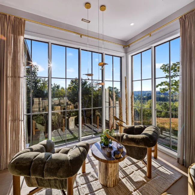 Corner breakfast nook with two olive-green tufted chairs, a round wood table and floor-to-ceiling black-framed windows over a view