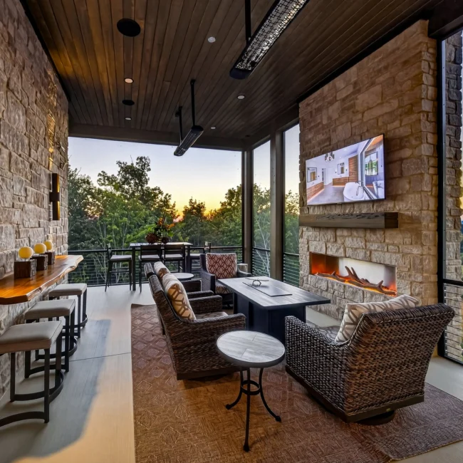 Sunset Heights covered porch at dusk with wicker chairs around a fire table, a stone fireplace with TV and a live-edge ledge