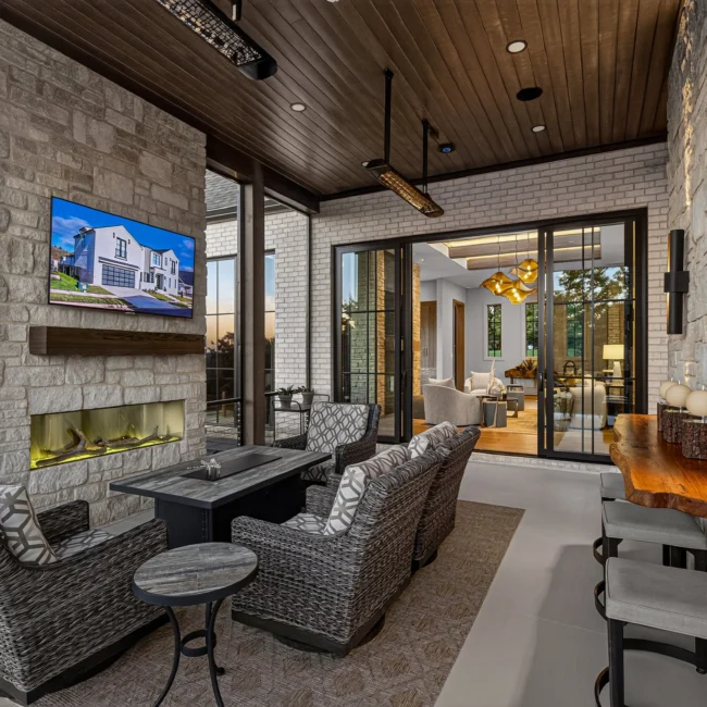 Covered porch seating area with wicker swivel chairs, a stone fireplace, a heater overhead and glass doors into the great room