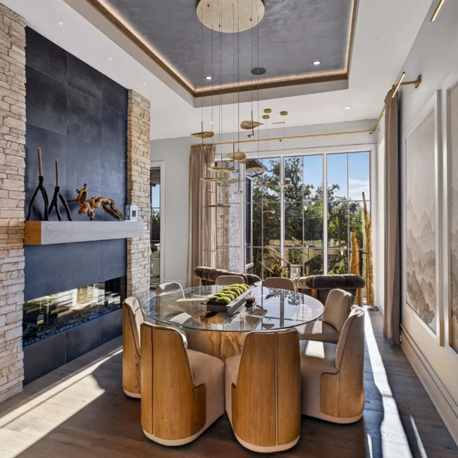 Sunset Heights round glass-top dining table with curved wood chairs in front of a two-sided fireplace under a dark plaster ceiling