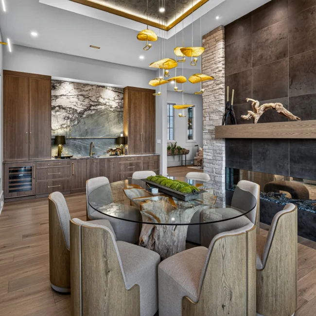 Sunset Heights round glass dining table on a driftwood base, with a stacked stone fireplace, a bar wall and gold leaf pendants