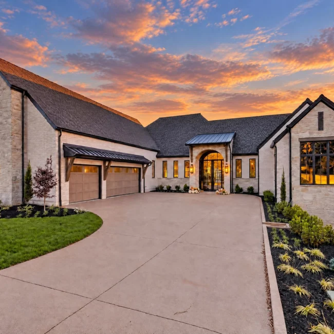 Sunset Heights front exterior at dusk with painted brick and stone, dark roof, arched entry and a curved concrete drive
