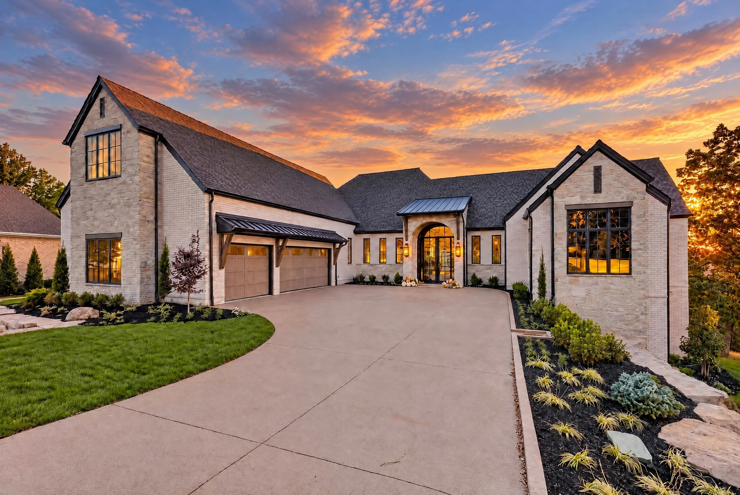 Sunset Heights front exterior at dusk with painted brick and stone, dark roof, arched entry and a curved concrete drive