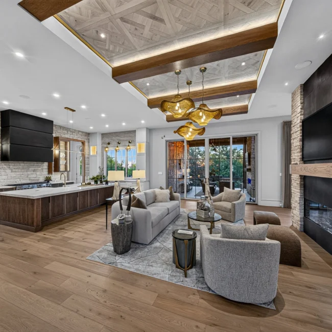 Sunset Heights great room with a stone fireplace column, gray sofas, a beamed parquet ceiling with gold pendants and an island kitchen