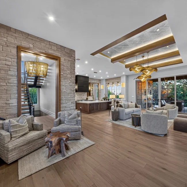 Wide view from the entry toward a great room with armchairs, a stone wall, a beamed ceiling and an open kitchen
