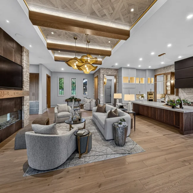 Long view along the great room with a stone fireplace, gray curved sofa, beamed ceiling and island kitchen beyond