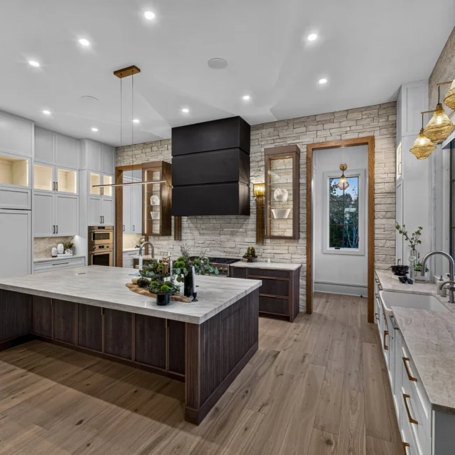 Sunset Heights kitchen at dusk with an island with a stone top, white cabinets, a black range hood and brass pendants