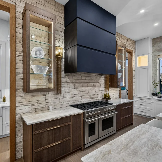Gas range with a black hood in stacked stone with a wood-framed cabinet, and the island counter edge in the foreground