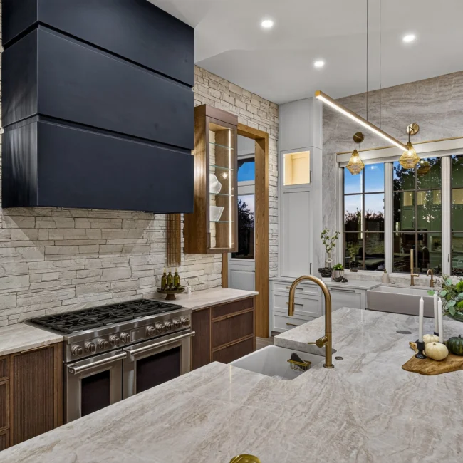 Kitchen range with a black hood, stacked stone wall and brass faucet at the island, with a window over the sink beyond