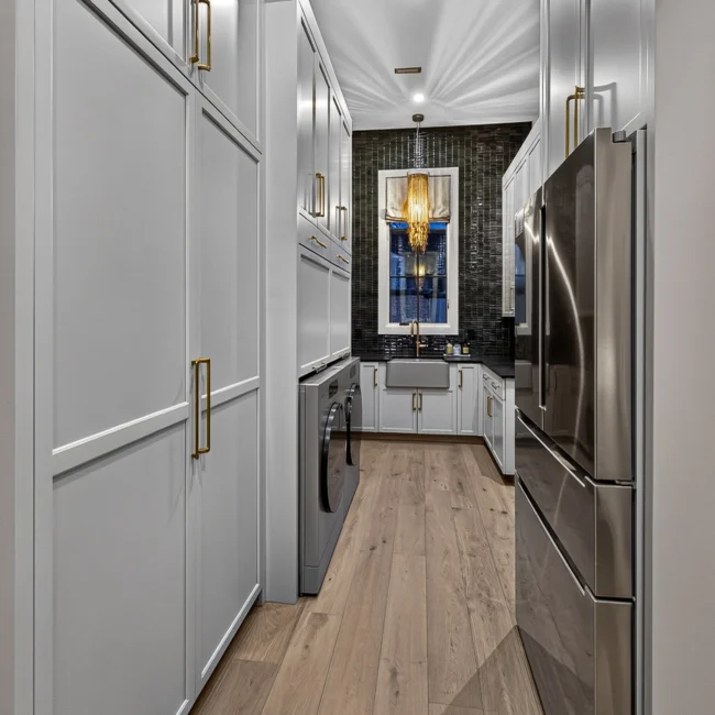 Utility hallway with white paneled cabinets and brass pulls, a washer and refrigerator, ending at a window with a chandelier