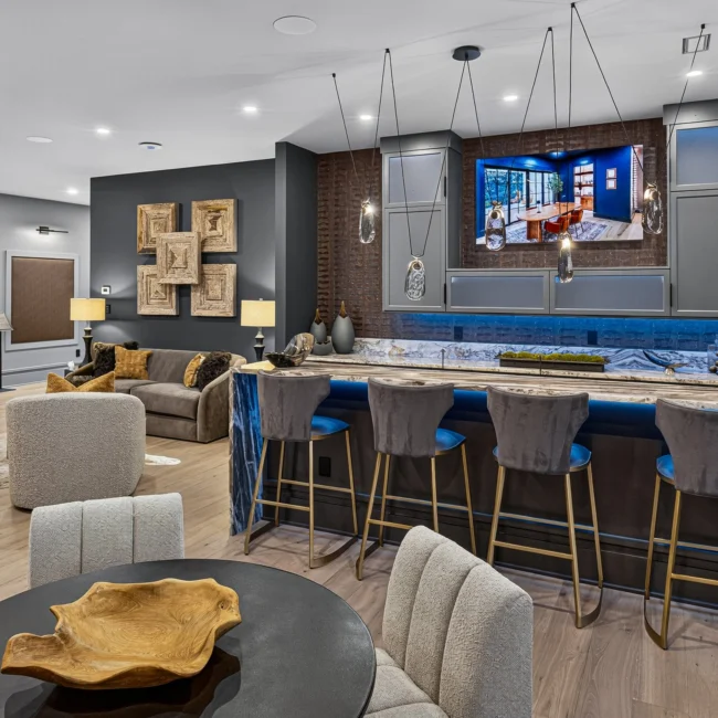 Lower-level bar with blue-backed stools along a stone waterfall counter, a TV in the cabinet wall and brass legs