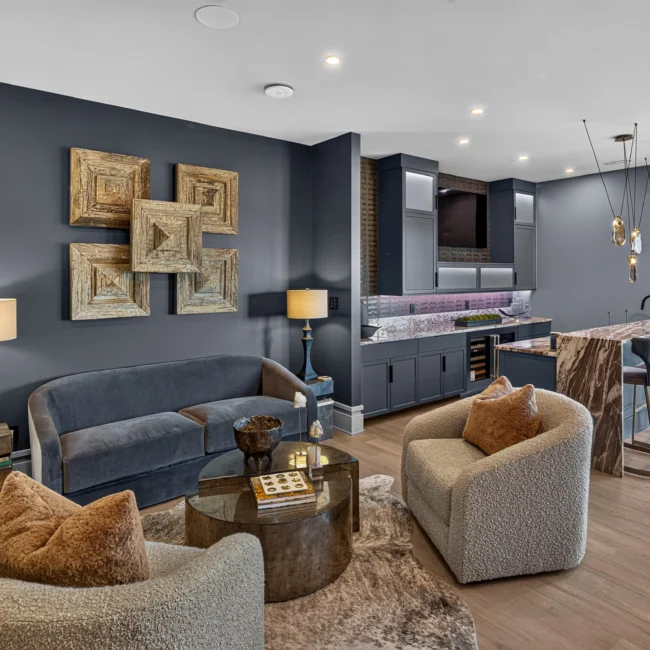 Lower-level lounge with a navy accent wall, velvet sofa, boucle chairs and a wet bar with a stone counter and four stools