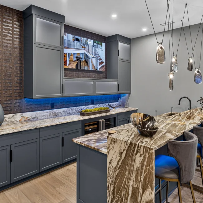 Sunset Heights wet bar with gray cabinets, a veined stone counter and waterfall edge, blue under-cabinet lighting and gray velvet stools