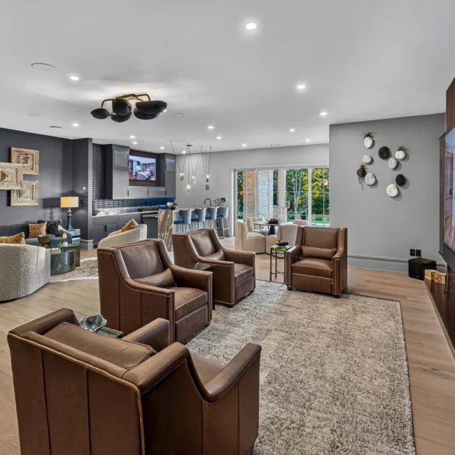 Lower-level media area with a wall-mounted TV on a wood-slat wall, four brown leather chairs and a bar beyond