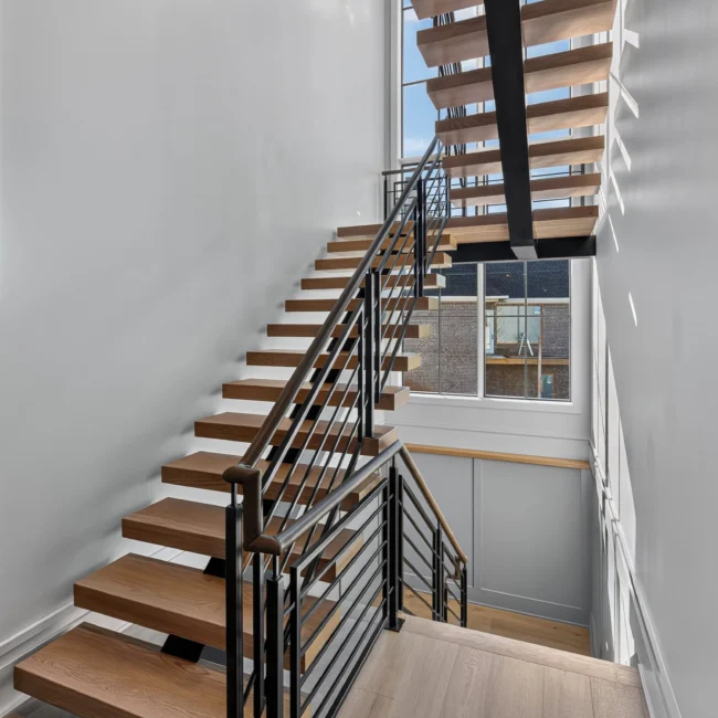 Open wood stair with floating treads and black steel railing, seen from below beside a tall window