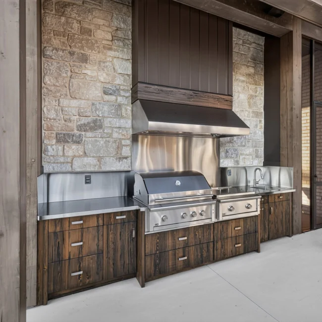 Outdoor kitchen with a stainless grill, a steel hood against stone and dark reclaimed wood drawer cabinets