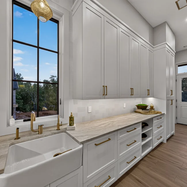 Pantry with a farmhouse sink beneath a tall window, white cabinets with brass pulls and a glass door to the outside