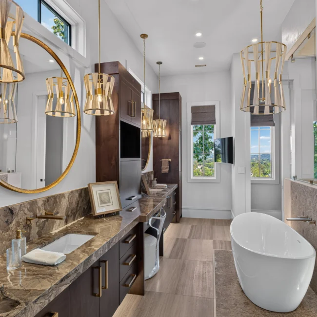 Primary bath with a long dark wood vanity, round brass mirror, three pendants and a white freestanding tub