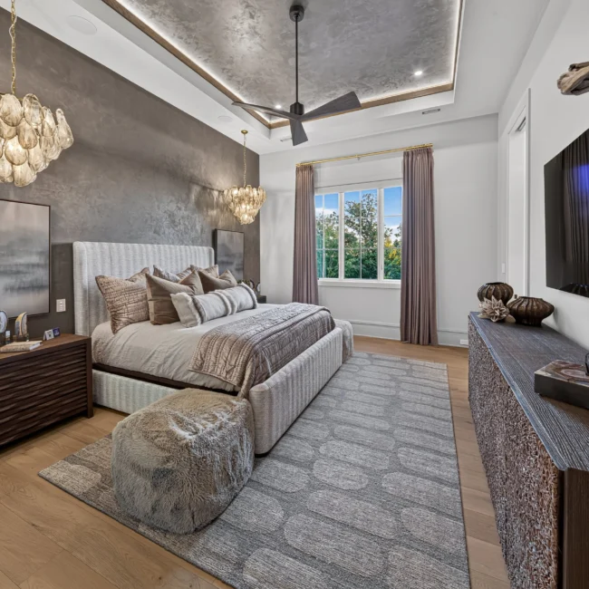 Primary bedroom by Mike Stevens Homes with a metallic plaster tray ceiling, upholstered bed, ceiling fan and chandelier beside the window
